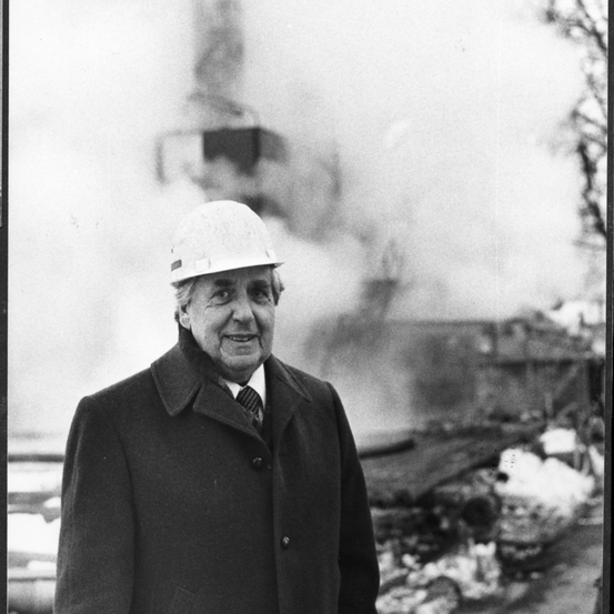Ein Mann in einer Schutzhelm und einem Mantel steht draußen, vor einem rauchigen Hintergrund mit Schutt und Trümmern.