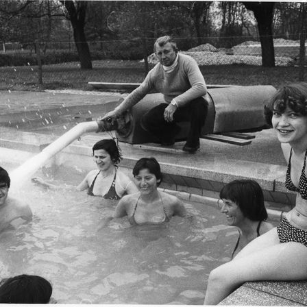 Ein Schwarz-Weiß-Foto von fünf Personen in einem Pool, eine sitzt auf einer Kante. Ein Mann mit einem Schlauch spritzt Wasser in den Pool. Bäume und ein Zaun im Hintergrund.