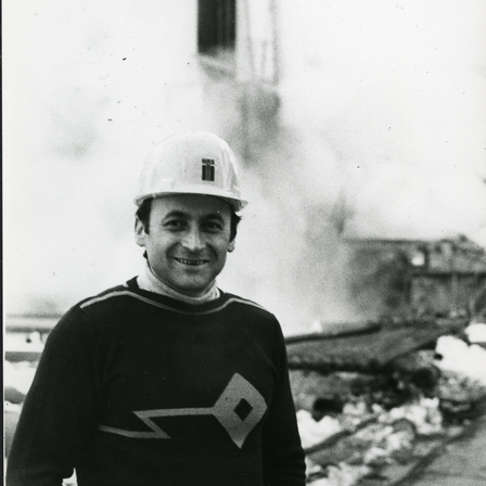 Ein Schwarz-Weiß-Foto zeigt einen Mann mit weißem Helm vor einer Baustelle.