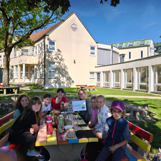 Eine Gruppe von Kindern und Erwachsenen sitzt an einem Picknicktisch in einem Park vor einem Gebäude. Sie lächeln und halten ein Zertifikat.