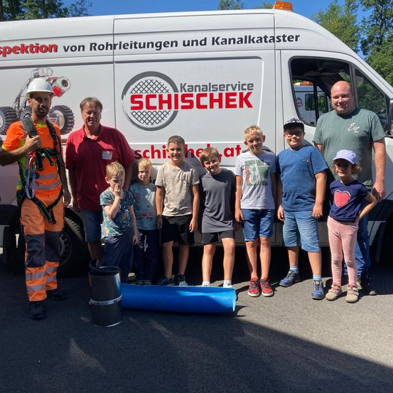 Erwachsene und Kinder posieren vor einem weißen Van mit 'Kanalservice SCHISCHEK' drauf. Einige Kinder halten eine Rolle blauen Stoffs.