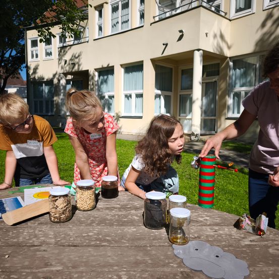 Drei Kinder und ein Erwachsener sind draußen und untersuchen Gläser mit verschiedenen Inhalten auf einem Picknicktisch, mit einem Gebäude im Hintergrund.