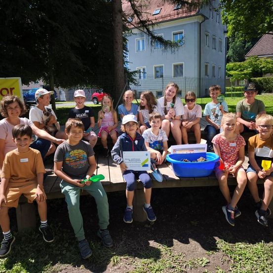 Eine Gruppe von Kindern und Erwachsenen sitzt auf einer Parkbank. Sie halten Lupen und einige tragen Mützen. Ein Kind hält ein Zertifikat. Bäume und ein Gebäude sind im Hintergrund.