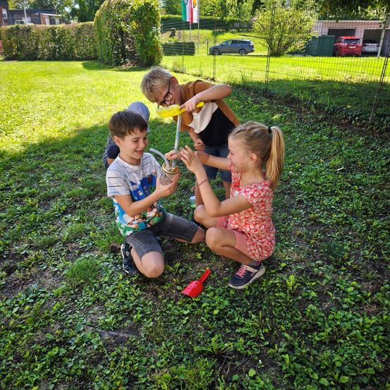 Drei Kinder spielen auf einem Rasen. Eines hält eine gelbe Gießkanne. Hinter ihnen ist ein Zaun, ein Busch und geparkte Autos.