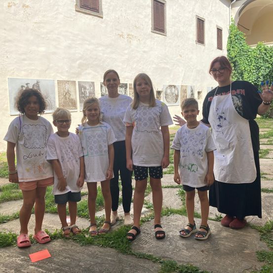 Eine Gruppe von Kindern und zwei Frauen steht vor einem weißen Gebäude, alle in weißen T-Shirts. Eine Frau trägt ein weißes Schürzenkleid mit einem schwarzen Shirt.
