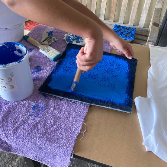 Eine Person malt eine blaue Schale auf einem braunen Tisch, mit einem Pinsel und einem Eimer blauer Farbe in der Nähe.