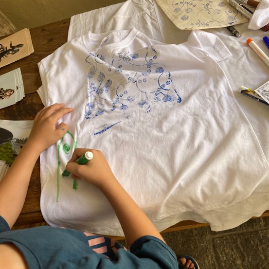 Ein Kind verziert ein weißes T-Shirt mit blauen Blumenmustern und grünen Linien. Der Schreibtisch ist mit Papieren, Stiften und Markern übersät.