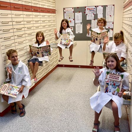 Sechs Kinder in Laborkittel sitzen auf Regalen und halten Bücher in den Händen. Sie scheinen für ein Foto in einer Apotheke zu posieren.