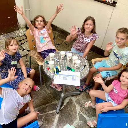 Eine Gruppe von Kindern sitzt um einen Tisch, lächelt und winkt. Ein Papier mit Schrift und Bechern ist auf dem Tisch. Sie tragen lässige Sommerkleidung.