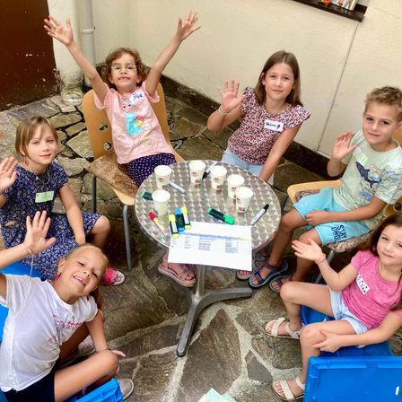 Eine Gruppe von Kindern sitzt um einen Tisch, lächelt und winkt. Ein Papier mit Schrift und Bechern ist auf dem Tisch. Sie tragen lässige Sommerkleidung.