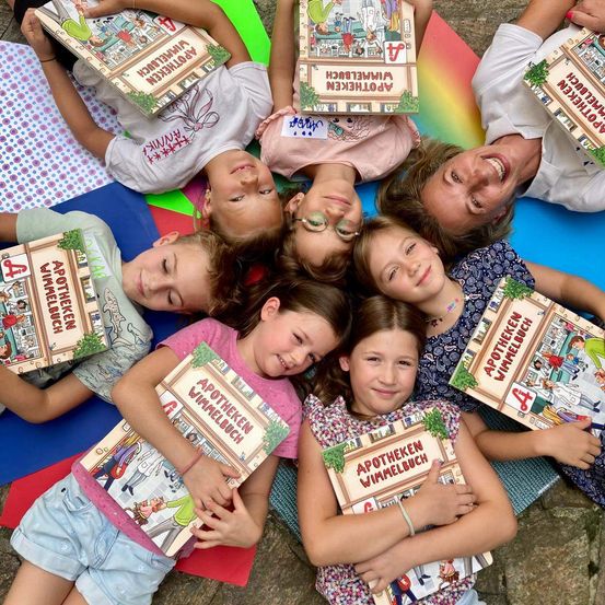 Mehrere Kinder liegen auf einer bunten Decke, jedes hält ein Buch mit dem Titel 'Apotheken Wimmelbuch'. Sie lächeln und posieren für ein Foto. Eine Frau steht hinter ihnen und lächelt. Das Bild ist aus einer Vogelperspektive aufgenommen.