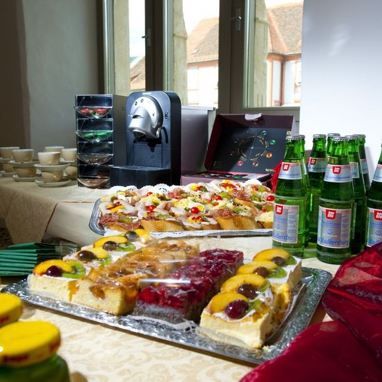 Ein Tisch mit verschiedenen Desserts, einschließlich Kuchen, Obsttörtchen und einer Kaffeemaschine. Flaschen grüner Limonade sind aufgereiht, und eine Schachtel Süßigkeiten steht auf dem Tisch. Draußen sind Gebäude durch ein Fenster sichtbar.