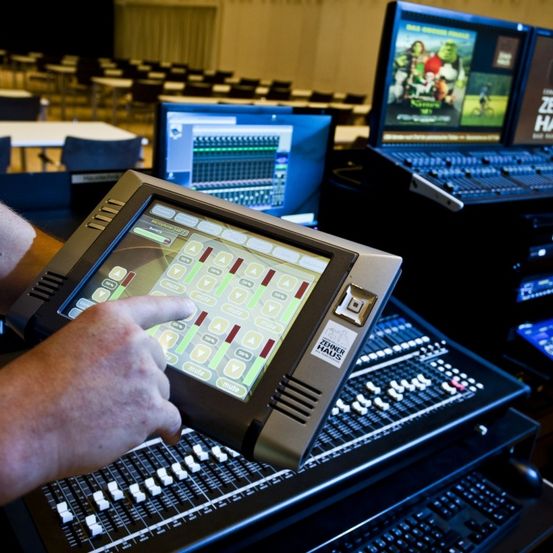 Eine Hand berührt einen Touchscreen auf einer Mischkonsole. Dahinter zeigen Bildschirme eine Filmszene, und der Raum ist mit leeren Stühlen gefüllt.