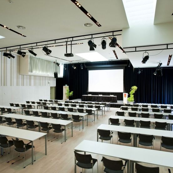 Ein leerer Konferenzraum mit Reihen von Tischen und Stühlen, einer großen Leinwand und an der Decke hängenden Scheinwerfern.