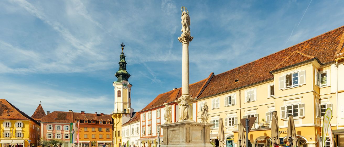 Ein Stadtplatz mit einer hohen Säulenstatue in der Mitte, umgeben von Gebäuden und Menschen. Es gibt geparkte Fahrräder an der Seite, und der Himmel ist blau mit Wolken.