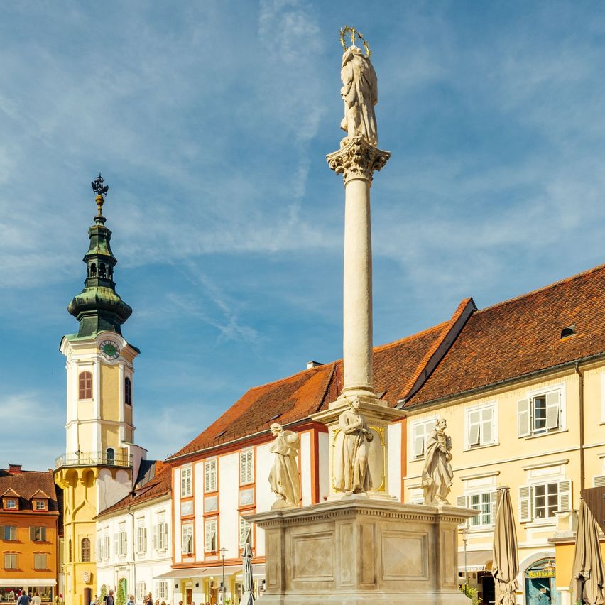 Ein Stadtplatz mit einer hohen Säulenstatue in der Mitte, umgeben von Gebäuden und Menschen. Es gibt geparkte Fahrräder an der Seite, und der Himmel ist blau mit Wolken.