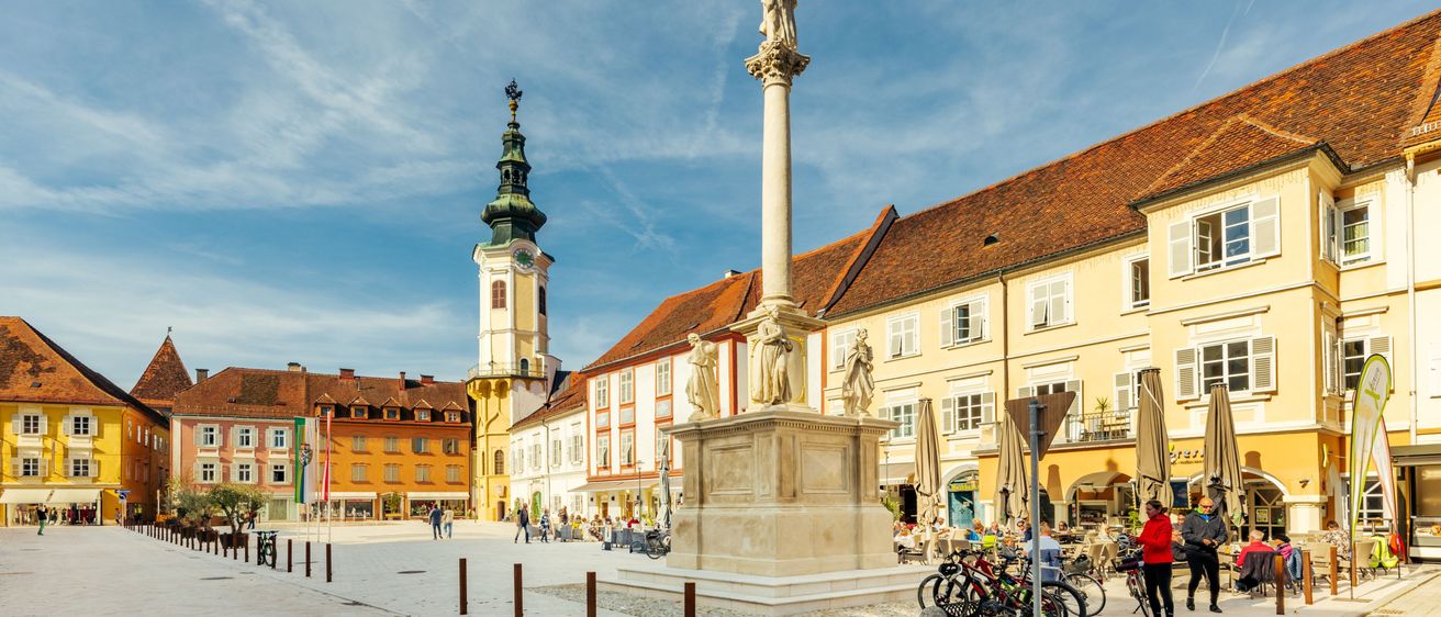 Ein Stadtplatz mit einer hohen Säulenstatue in der Mitte, umgeben von Gebäuden und Menschen. Es gibt geparkte Fahrräder an der Seite, und der Himmel ist blau mit Wolken.