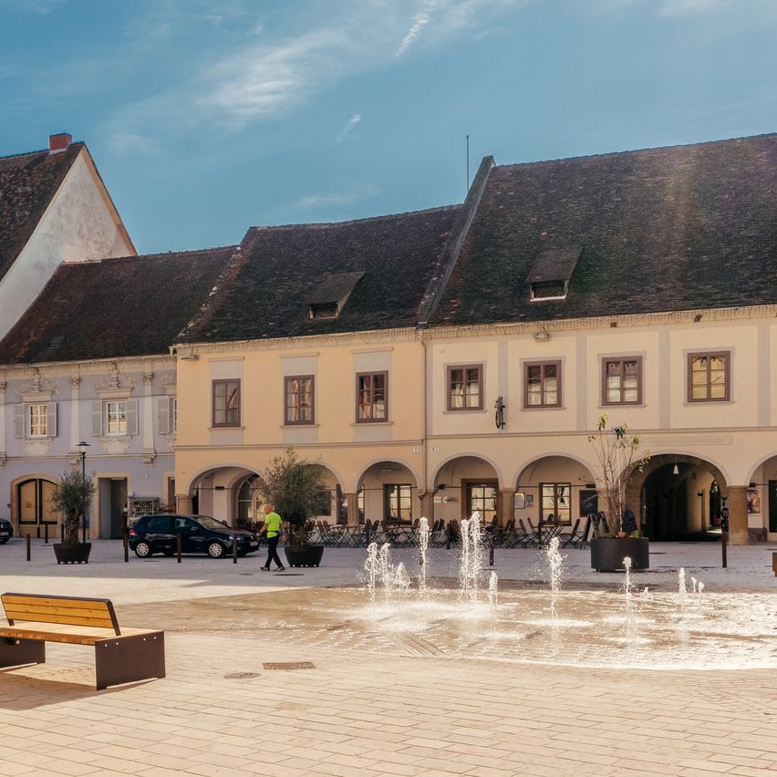Ein Stadtplatz mit einem Brunnen, umgeben von Gebäuden. Die Gebäude haben braune Dächer und weiße Wände. Eine Person läuft auf dem Bürgersteig.