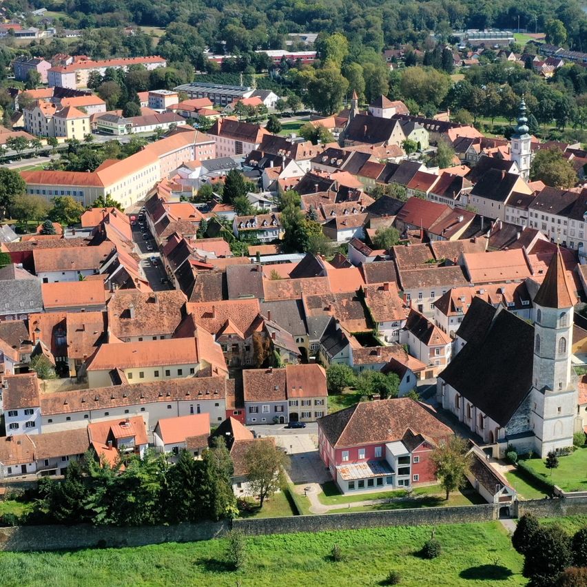 Luftaufnahme einer Stadt mit vielen Gebäuden, einige mit roten Dächern, umgeben von Bäumen und einem Fluss. Im Vordergrund ist ein grünes Feld.