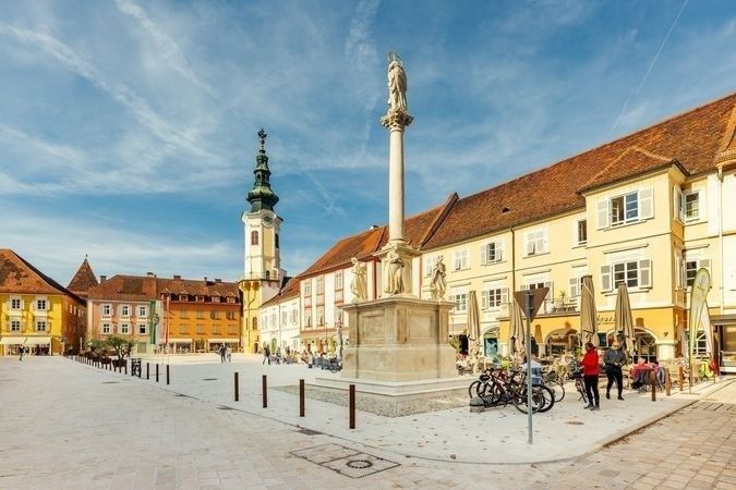 Ein Platz im Freien mit einer Statue und Radfahrern, umgeben von Gebäuden und Menschen, die an einem sonnigen Tag spazieren.
