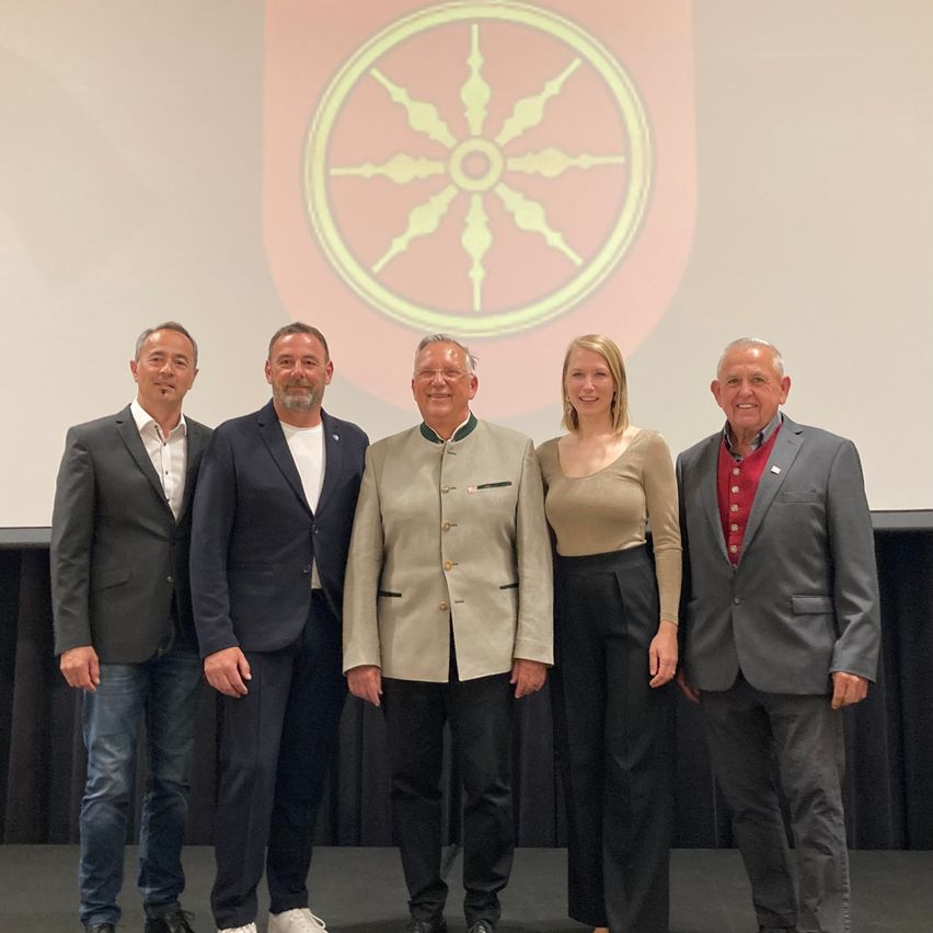 Fünf Personen stehen lächelnd auf einer Bühne für ein Foto. Hinter ihnen zeigt eine Leinwandprojektion ein großes kreisförmiges Design mit einem Symbol.