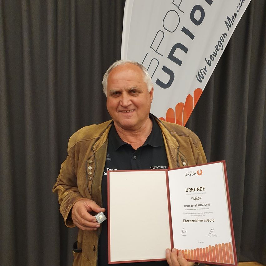 Ein Mann hält einen Zertifikat in der Hand, lächelt und steht vor einem Hintergrund mit einem Banner, auf dem 'Sport Union' steht. Das Zertifikat hat eine goldene Auszeichnung.