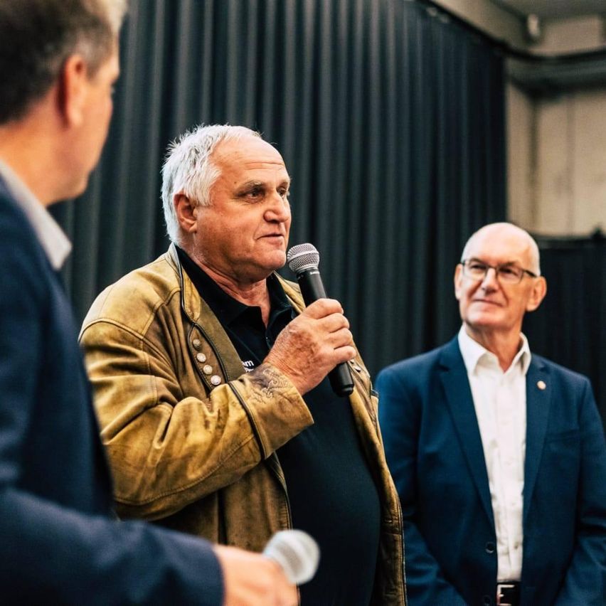 Ein älterer Mann spricht in ein Mikrofon in einem Konferenzraum. Hinter ihm stehen zwei andere Männer, einer in Anzug und der andere in einer Lederjacke.