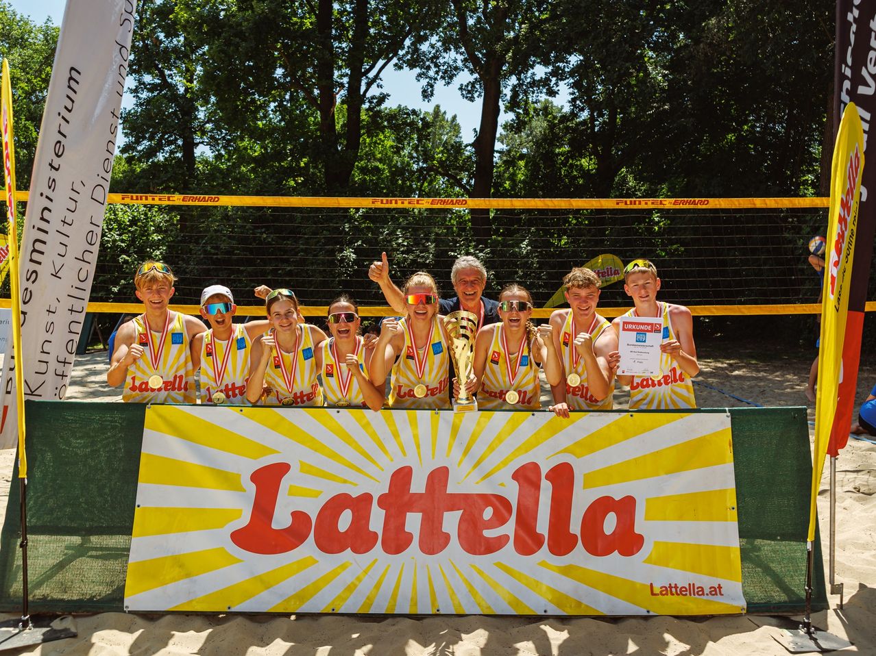 Eine Gruppe junger Athleten in gelben Trikots posiert für ein Foto am Strand, hält Medaillen und einen Pokal und hat ein Latella-Banner davor.