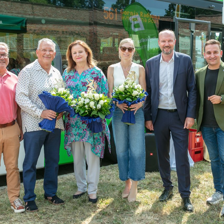 Fünf Menschen stehen vor einem Bus mit einem grünen Banner und der Nummer 561. Sie halten Blumen und lächeln. Der Bus ist auf Gras geparkt.