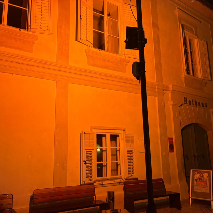Ein altes Gebäude mit gelben Wänden und weißen Fensterläden. Zwei Holzbänke stehen davor. Ein Schild ist auf der rechten Seite platziert.