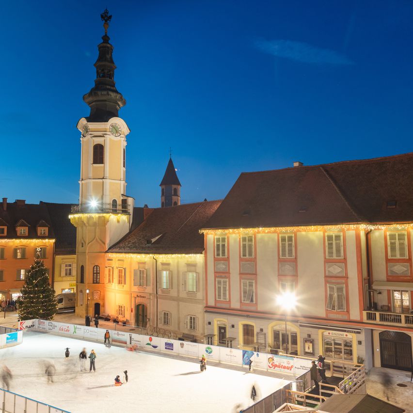 Eine Nachtansicht eines Stadtplatzes mit einer Eislaufbahn. Die Bahn ist von Gebäuden mit Weihnachtsdekoration umgeben. Ein Uhrturm ist im Hintergrund sichtbar.