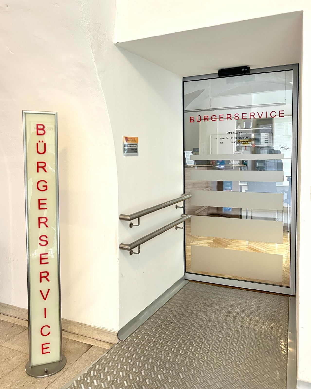 Ein Eingang mit einer Glastür, auf der 'BURGERSERVICE' steht. Die Tür ist offen und es gibt einen glänzenden Metallhandlauf. Der Raum im Inneren ist schwach beleuchtet.