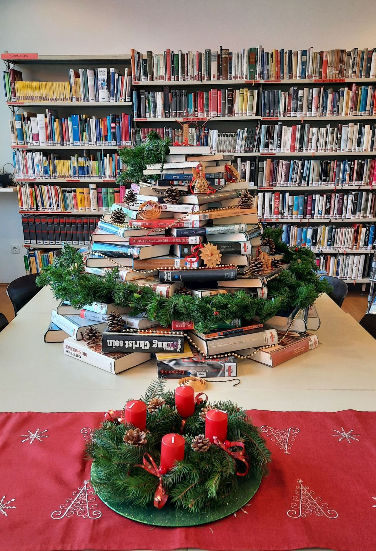 Ein Tisch mit einem Stapel Büchern, der in einer dreieckigen Form angeordnet ist. Die Bücher sind mit Tannenzapfen und Weihnachtsdekorationen verziert. Der Tisch ist mit einer roten Tischdecke bedeckt. Dahinter befinden sich mit Büchern gefüllte Regale.