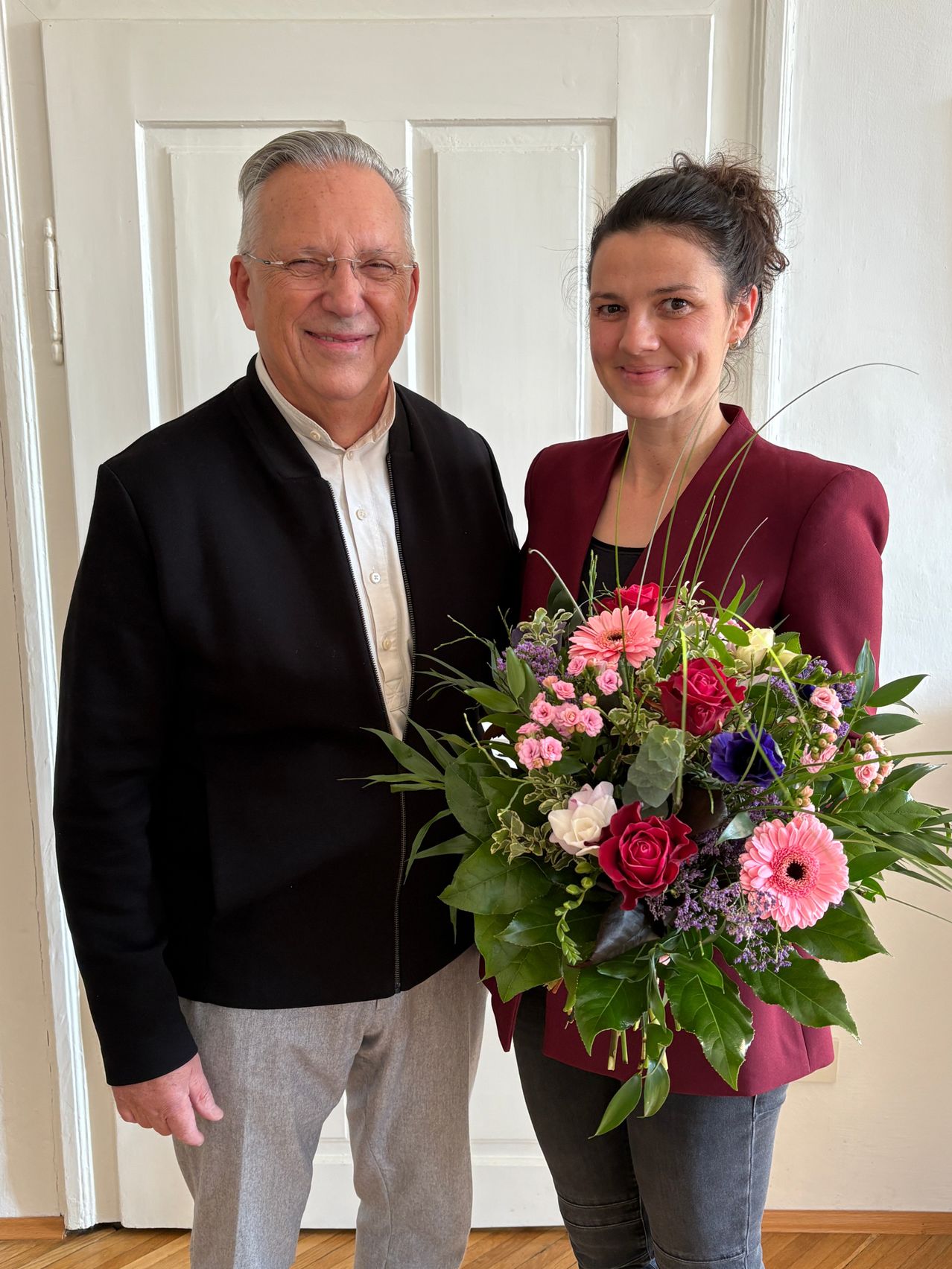 Ein älterer Mann und eine Frau stehen lächelnd vor einer weißen Tür und halten einen Blumenstrauß.
