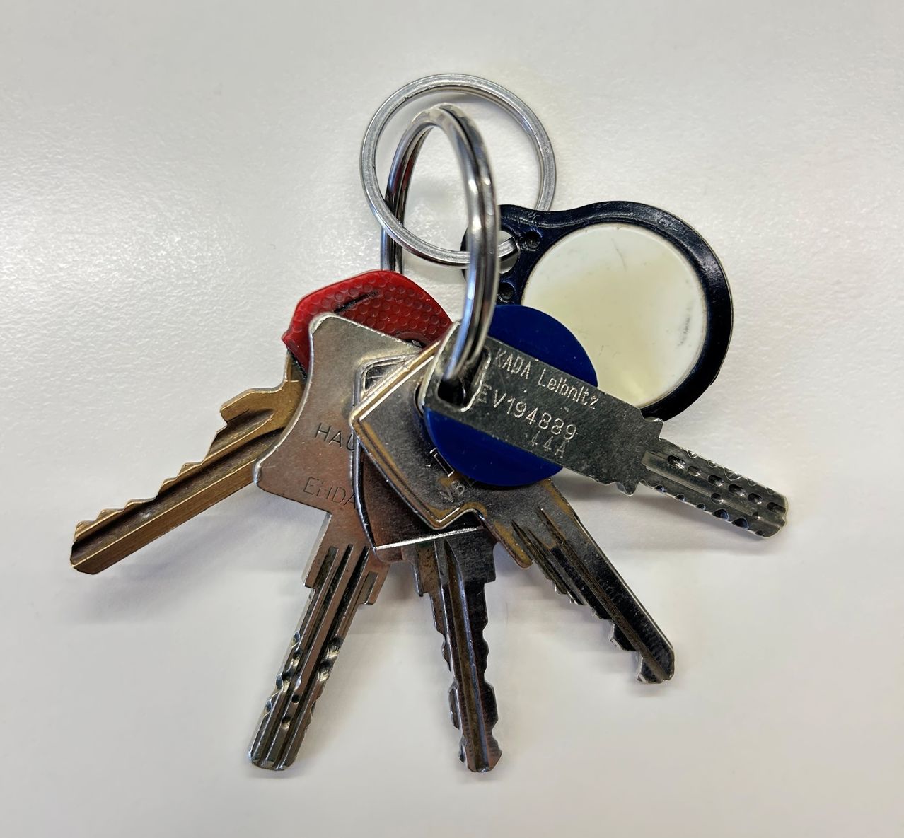 Ein Schlüsselbund mit mehreren Schlüsseln und einem kleinen Spiegel. Die Schlüssel haben verschiedene Größen und Farben, mit Etiketten wie 'KADA Lebnitz' und 'EV194889'.