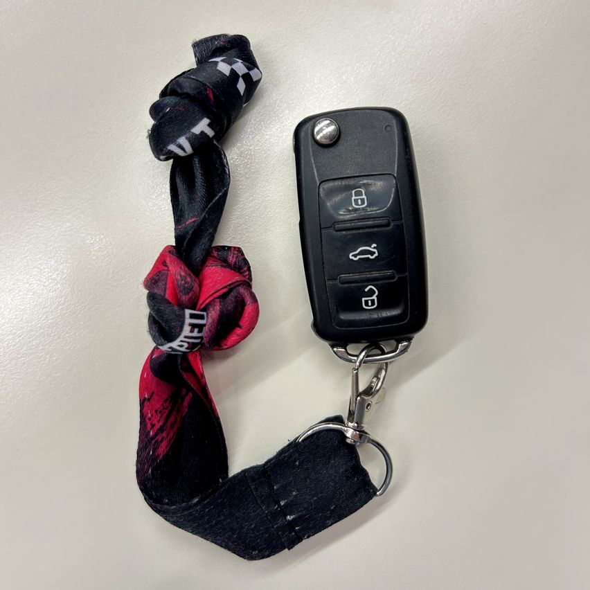 Ein schwarzer Autoschlüssel mit Schlosssymbol, Auto-Symbol und Schlüsselsymbol, befestigt an einem Jeansriemen mit rot-schwarzem Bandana.