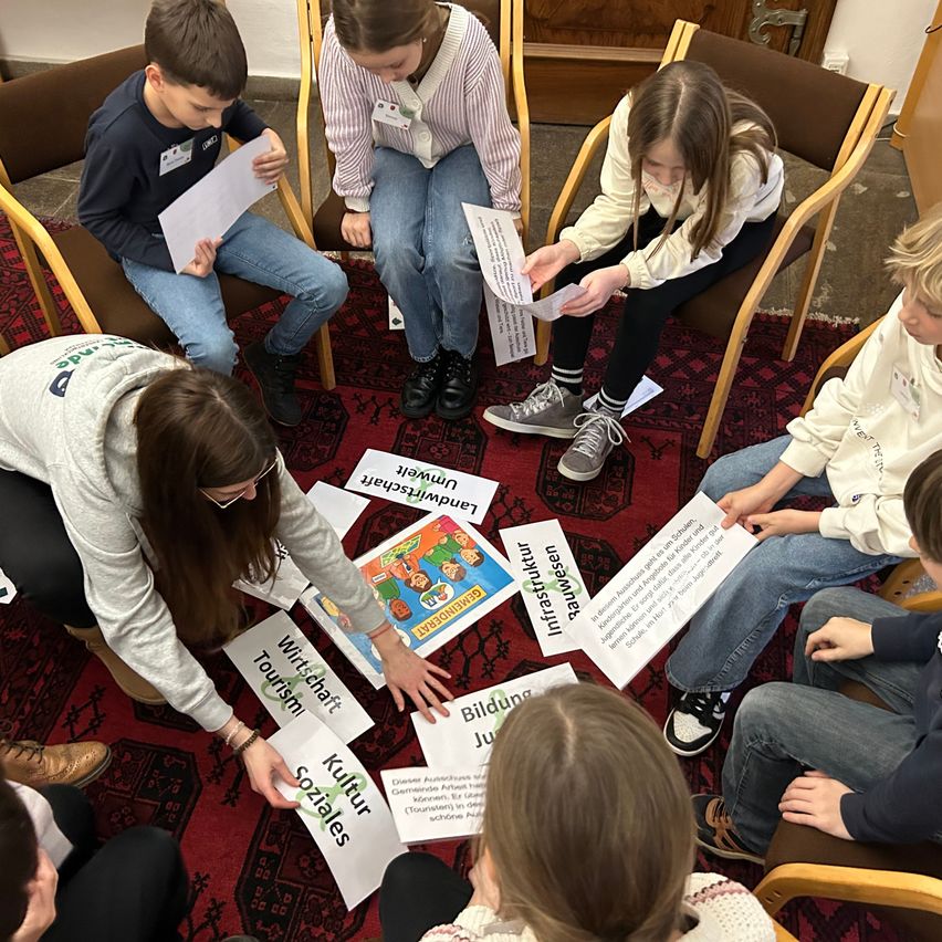 Eine Gruppe von Kindern und einer Frau sitzen im Kreis auf einem roten Teppich. Sie halten Papiere in den Händen und diskutieren etwas. Die Frau hält ein Buch. Die Papiere auf dem Teppich haben verschiedene Wörter darauf geschrieben.