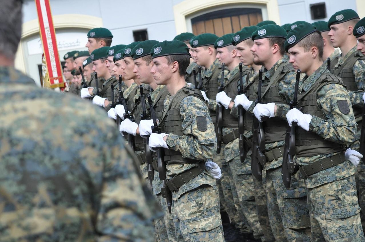 A group of soldiers in green berets and camouflage uniforms stand in a line with rifles. They are holding their rifles with white gloves and are probably in a parade.