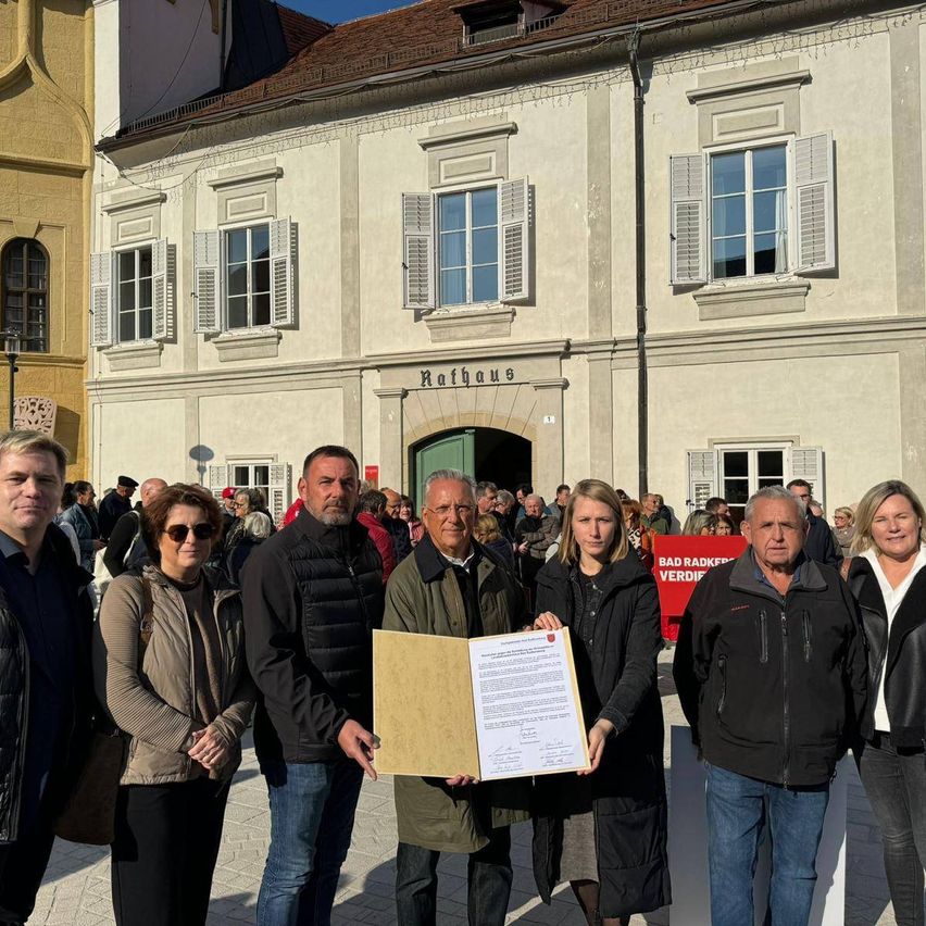A group of people stand in front of a building labeled Rathaus, holding a document. They are all dressed in winter clothes. The building has multiple windows with white shutters.