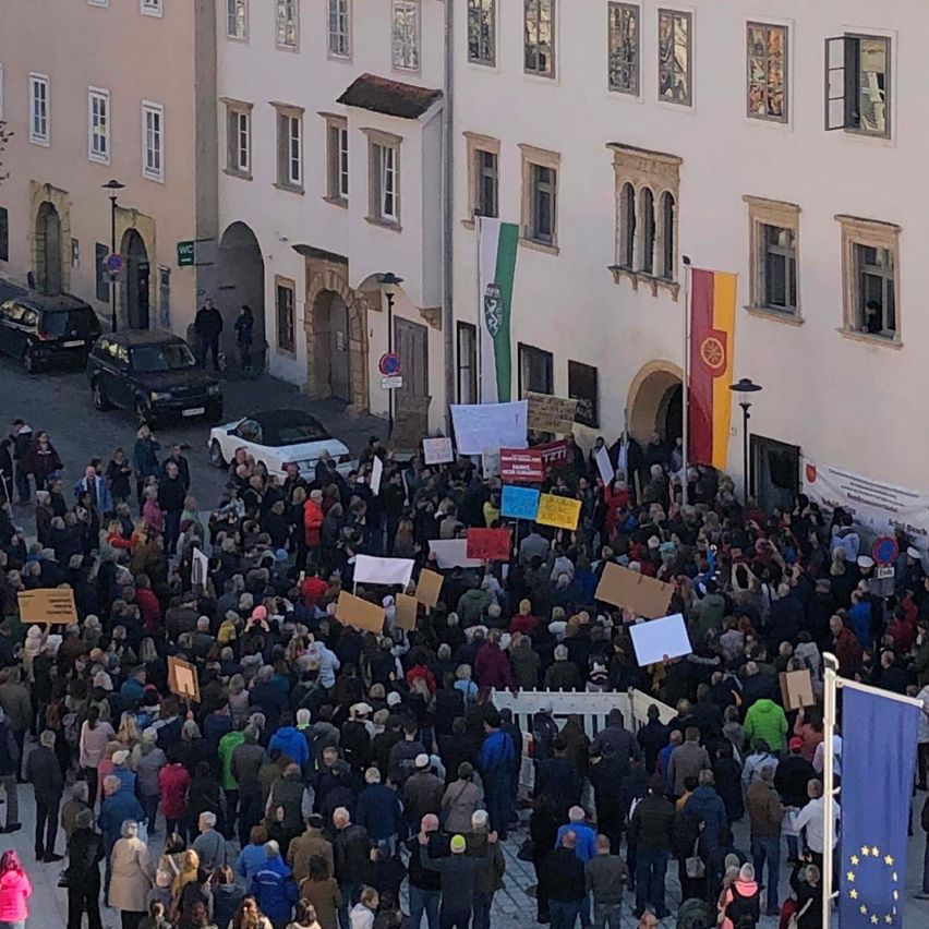 A crowd of people gathered in an outdoor area, holding signs and banners. The scene includes several cars parked on the street, a building with many windows, and a European flag on a pole.