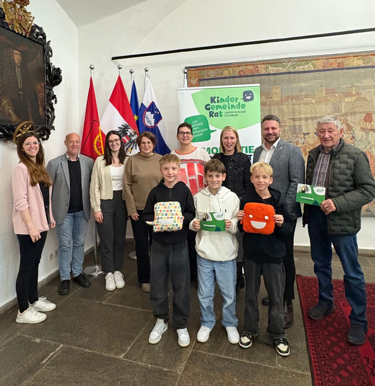 A group of adults and children pose for a picture in a room with flags and a banner. The children hold green items.