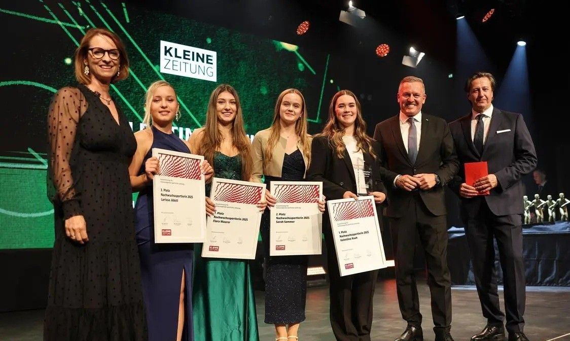 Five women and two men stand on stage, holding certificates, smiling. The backdrop features 'Kleine Zeitung' and green geometric shapes. Two men wear suits and ties, and the women wear dresses. The women hold certificates with names and positions.