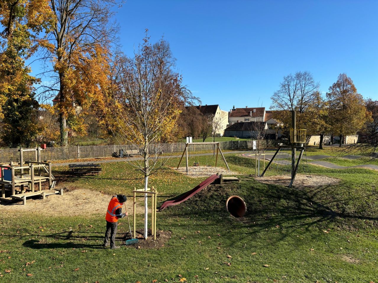Ein Arbeiter in einer orangefarbenen Weste pflanzt einen Baum in einem Park. Hinter ihm sind ein Rutschen und Spielgeräte zu sehen. Der Park ist von Bäumen und Gebäuden umgeben.