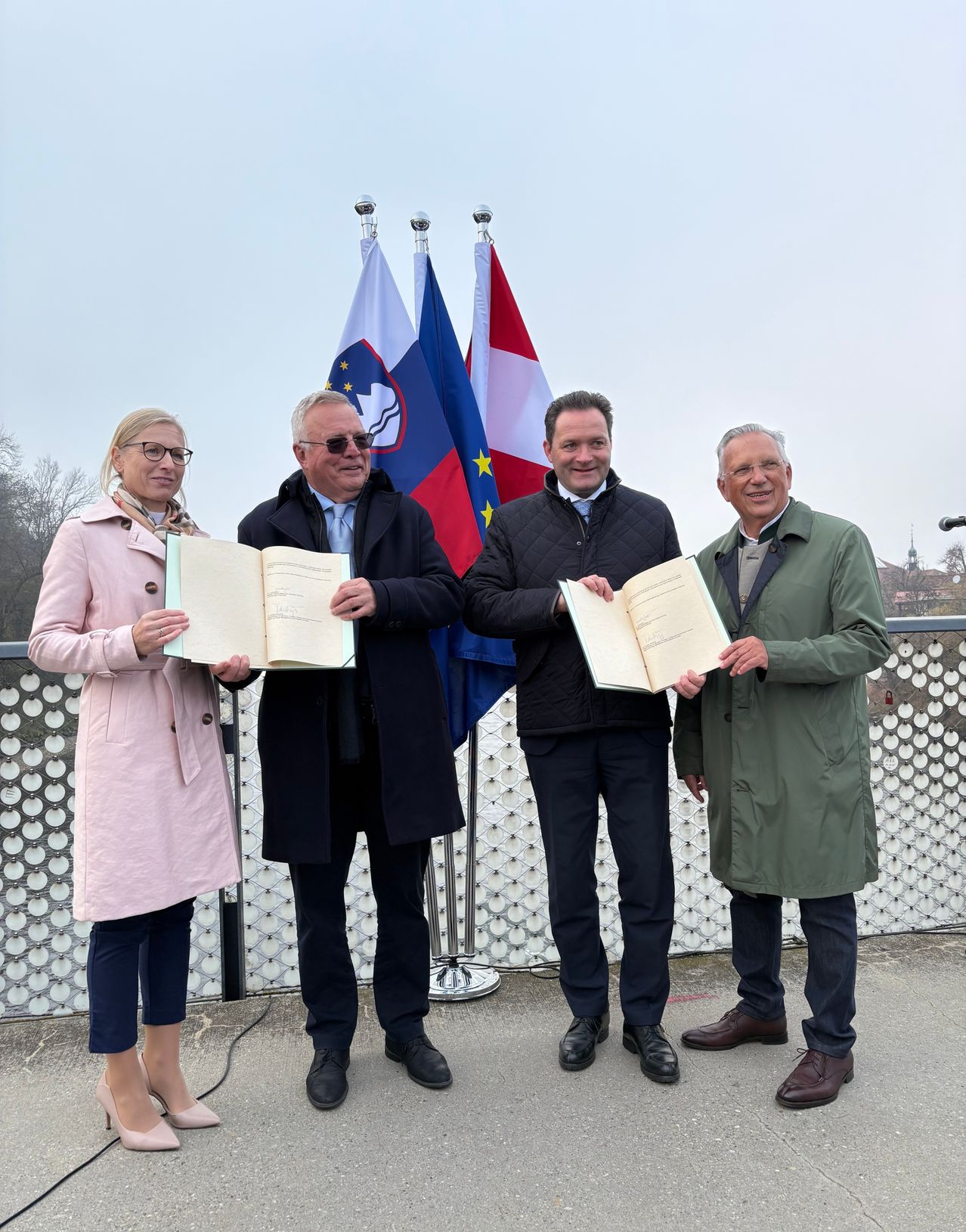 Four individuals stand in a formal pose with documents, against a backdrop of three flags, including the EU flag.