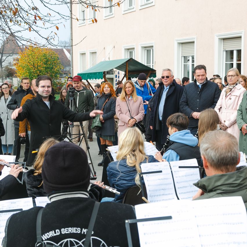 A group of people is gathered outside, likely for a musical performance. The man in the center is gesturing, and musicians with sheet music are seated in front. Spectators are standing and watching. The setting is in front of a building with trees and a canopy.