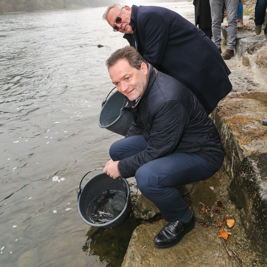 Two men are on the riverbank, one kneeling holding a bucket of fish, the other standing and looking at the fish.