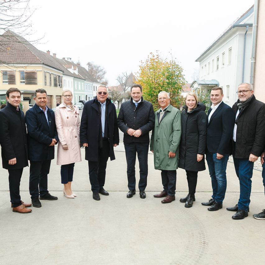 A group of adults stand together in a row, dressed warmly in coats and shoes, in front of buildings.