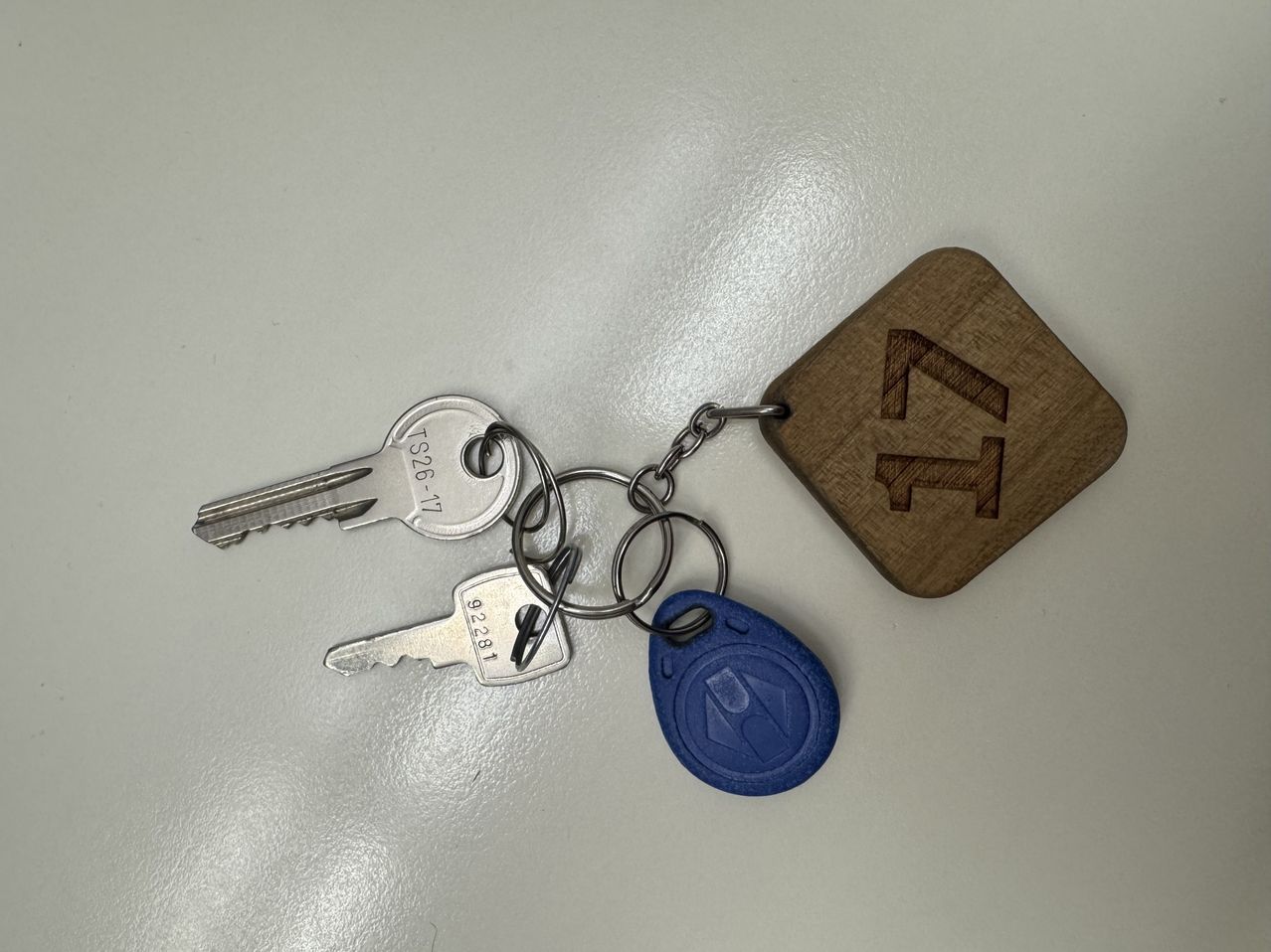Zwei Schlüssel und eine Zugangskarte mit einem blauen Tag mit der Nummer 47 liegen auf einer weißen Oberfläche. Die Schlüssel sind silber und die Karte ist blau.