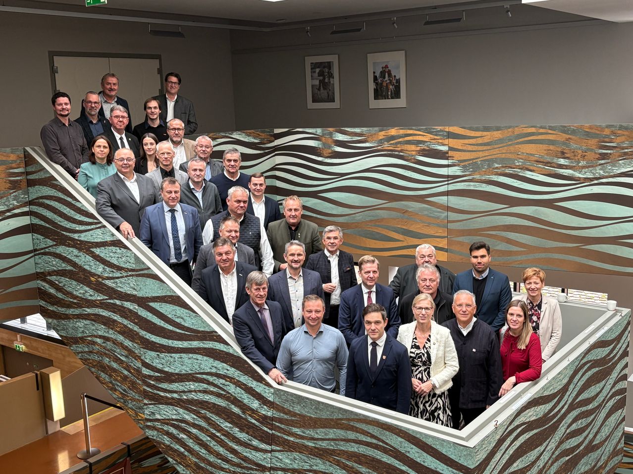 Eine Gruppe formell gekleideter Personen posiert auf einer Treppe. Dahinter hat eine Wand ein wellenförmiges Musterdesign und zwei gerahmte Bilder.