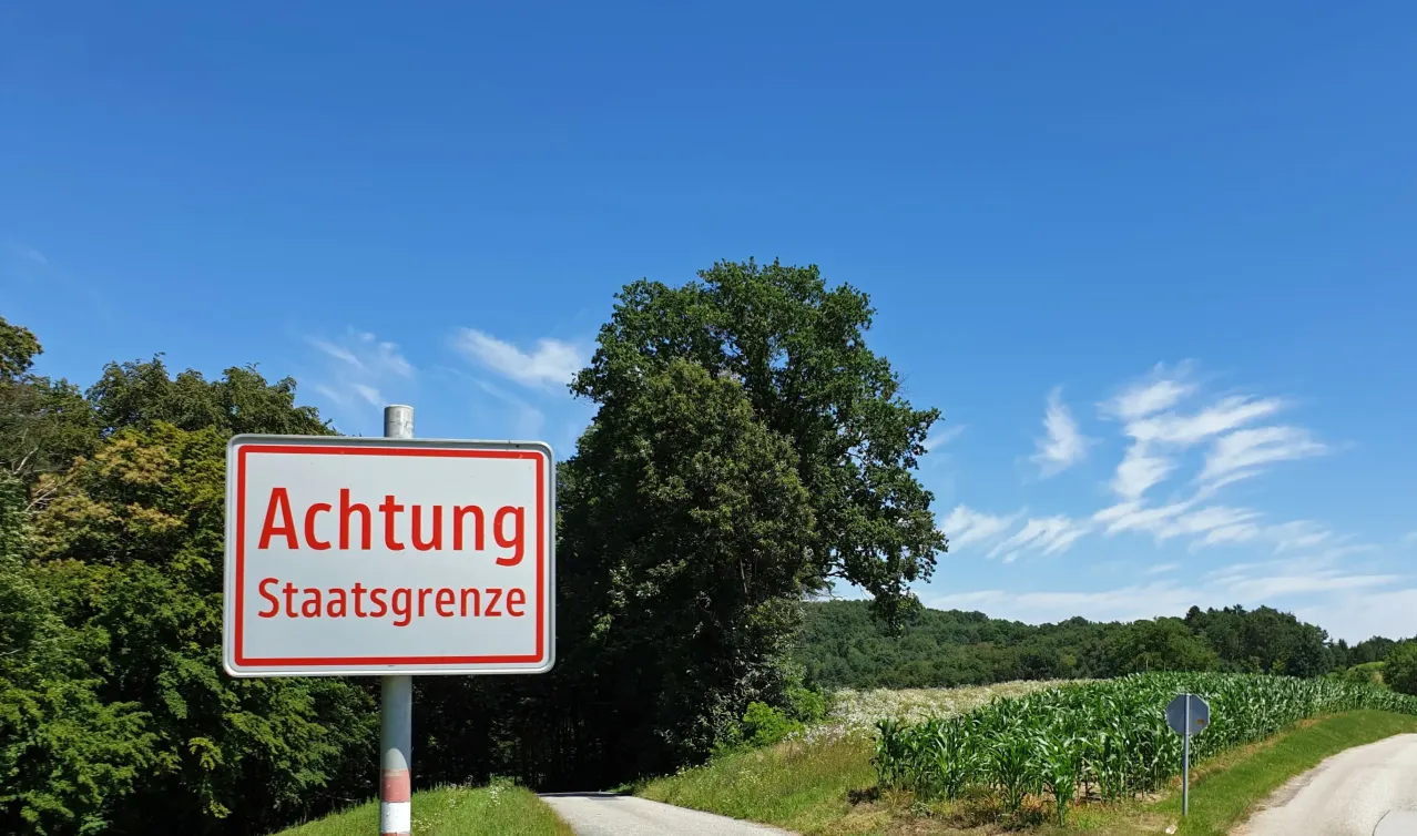 Ein Schild mit dem Wort 'Achtung' steht auf einem Pfosten am Straßenrand. Im Hintergrund befinden sich ein Maisfeld und ein Baum.